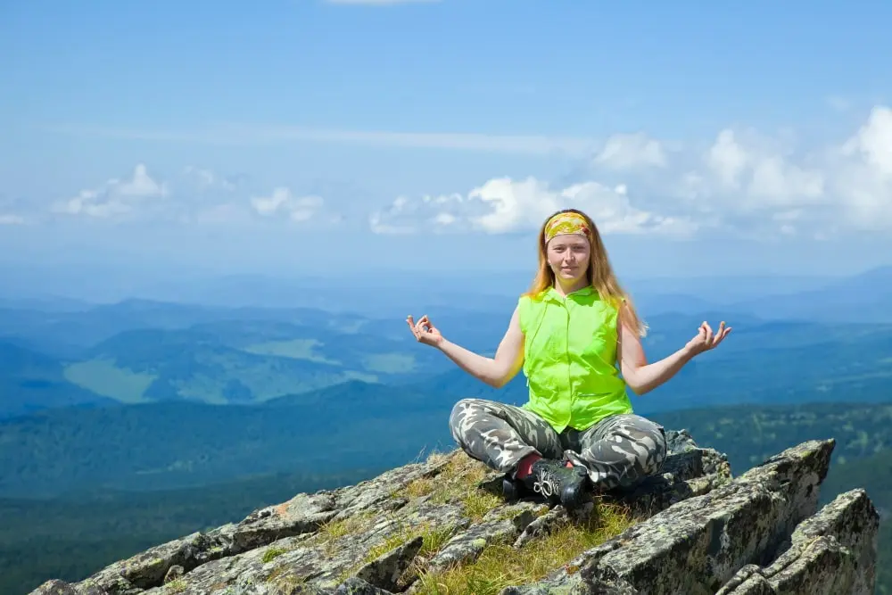 A woman sits cross-legged in a meditative pose on a rocky mountain peak.