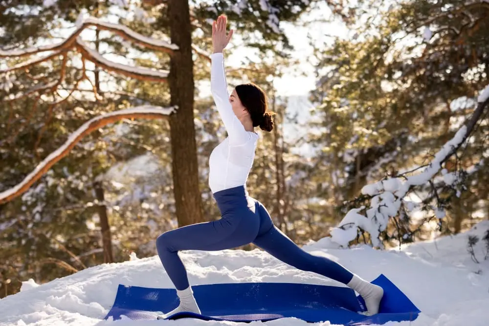 A woman doing yoga in the snow, standing in warrior pose on a blue yoga mat.