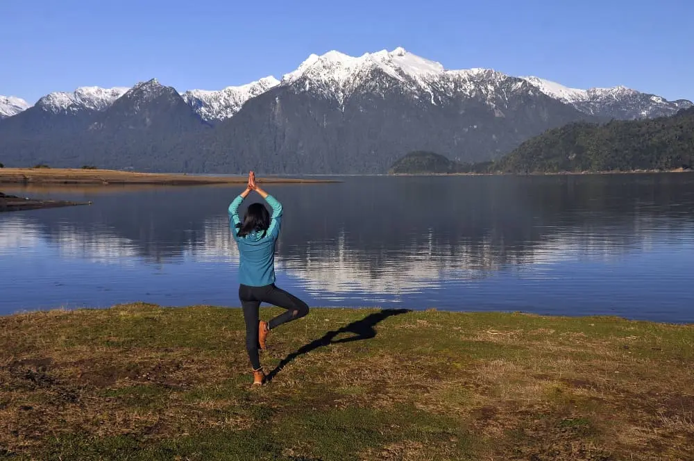 A man stands in a yoga tree pose on a grassy area by a tranquil lake.