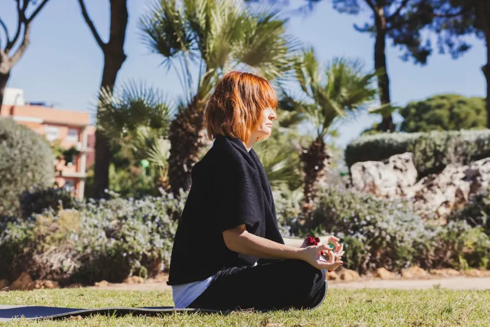A woman sits cross-legged on a grassy field and practices meditation in a peaceful outdoor setting.