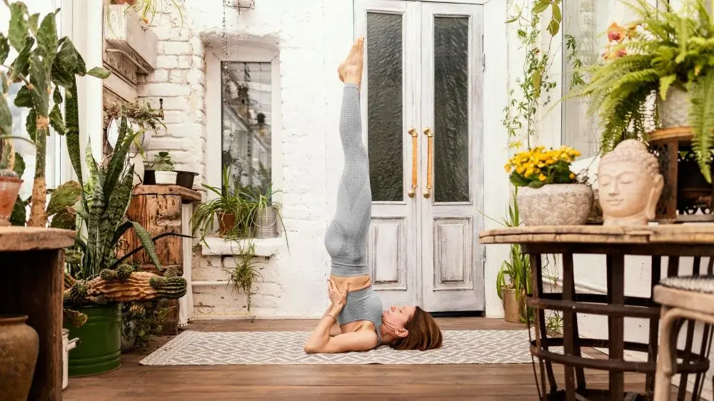 Woman performing yoga headstand pose indoors surrounded by plants