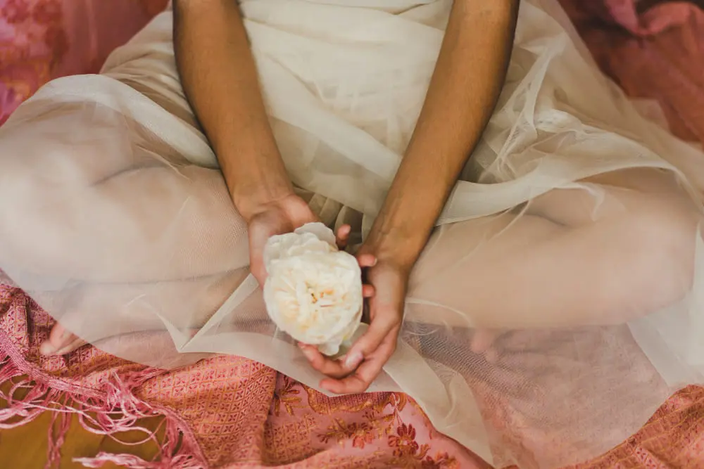 Woman in flowing white dress sitting cross-legged while holding a white peony flower