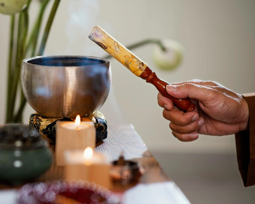 Hand striking a singing bowl with a mallet near candles, used in meditation or spiritual rituals.