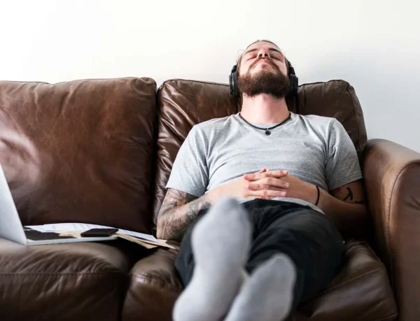 A man is relaxing on a brown leather couch, wearing headphones with his eyes closed. He has his hands clasped and is resting with his feet up, with a laptop and phone nearby. 