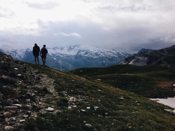 Two people are walking along a trail in the mountains, with snow-capped peaks in the background. The sky is overcast, and the landscape is lush with greenery and rocks. 