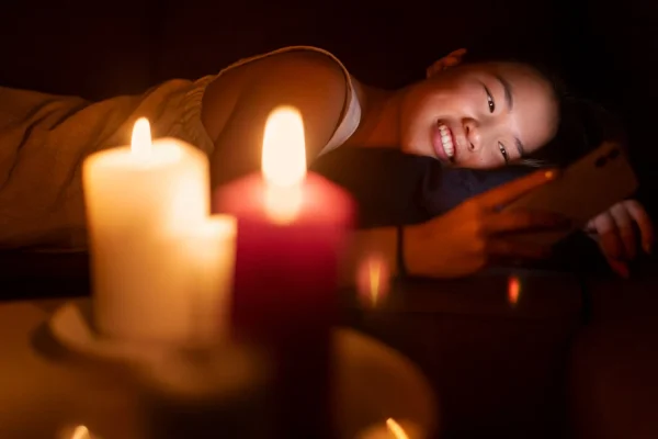 A woman relaxing on a couch by candlelight, smiling while looking at her phone in a cozy, warm atmosphere during the evening.